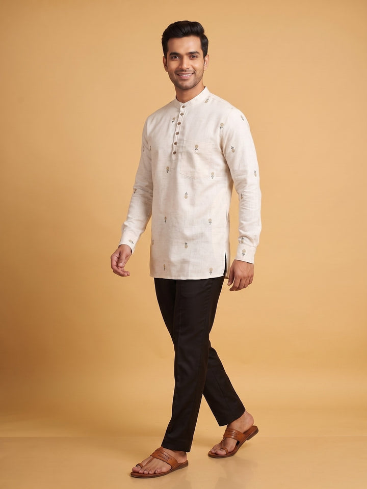 A man modeling a cream-colored long-sleeve kurta with small embroidered details, paired with black trousers and brown sandals, against a solid warm beige background. A man standing in an off-white cotton short kurta with minimalist floral embroidery, showcasing a relaxed fit.