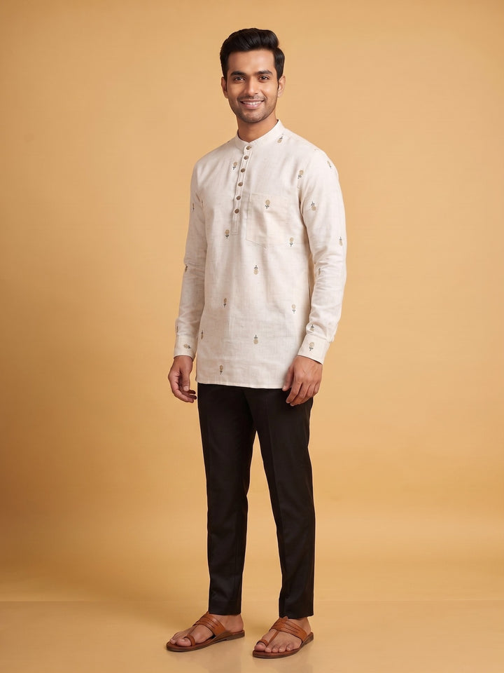 A man modeling a light beige long-sleeve kurta with small embroidered details, paired with black trousers and brown sandals, against a solid warm beige background. Full-length front view of a man modeling an off-white cotton short kurta featuring delicate floral embroidery and black pants.