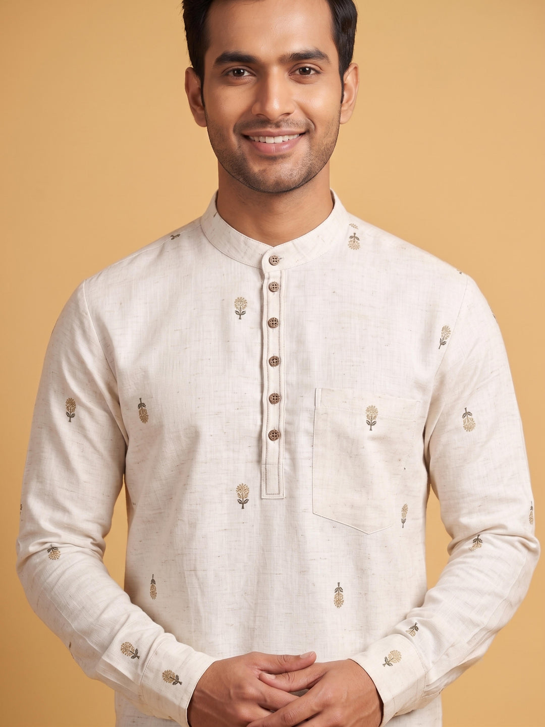 A man wearing a light beige linen kurta with small embroidered motifs and a buttoned placket, standing against a solid warm beige background. Medium shot of a man wearing an off-white cotton short kurta with minimalist floral embroidery and buttoned placket.
