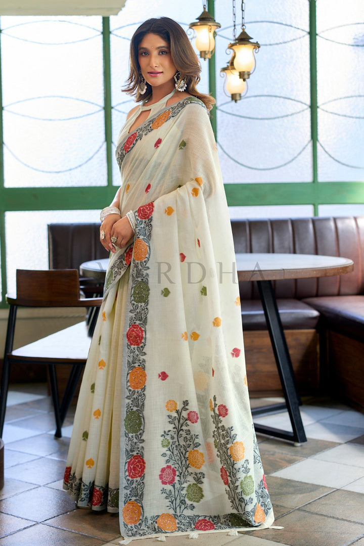 Woman wearing a floral saree in an indoor setting Side profile of a woman draped in an off-white linen saree, highlighting the striking charcoal-based floral jacquard border and elegant drape.