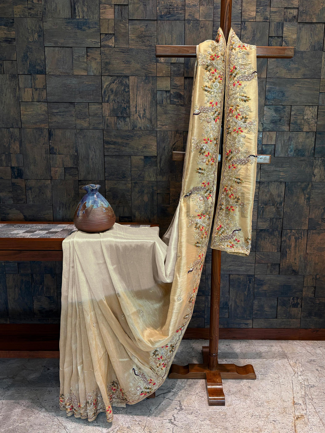 Off white tissue silk saree draped with multicolor hand zardozi embroidery and scalloped border on golden zari weave. Decorative fabric with floral patterns draped over a stand against a wooden paneled wall.
