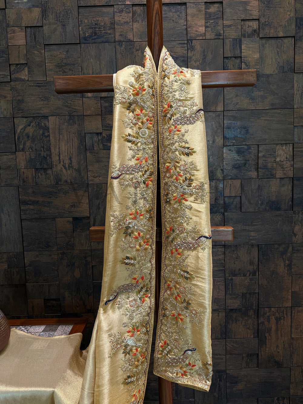 Embroidered scalloped border view with antique gold zari detailing and fine beadwork on off white silk. Decorative robe with floral patterns on a wooden cross against a textured wall.