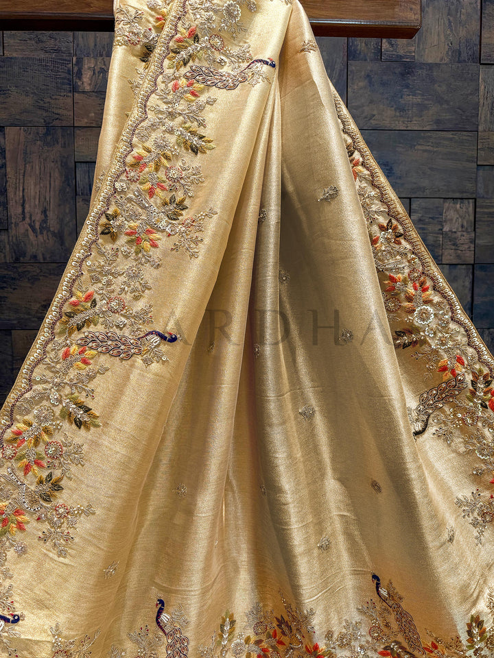 Pallu close-up showing detailed peacock motifs in red, orange, and green thread with metallic and sequin highlights. Beige fabric with intricate embroidery and peacock designs on a wooden surface.