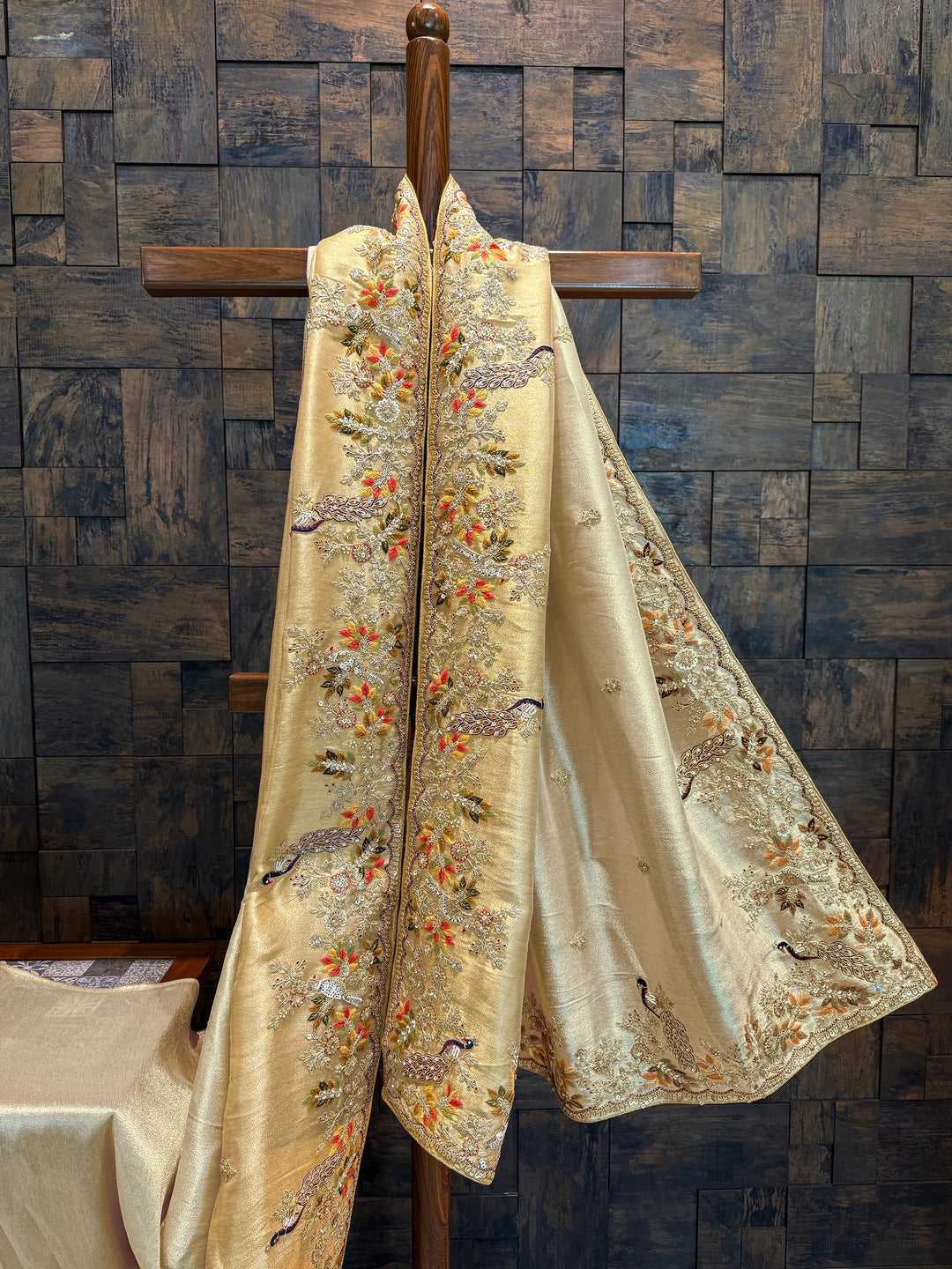 Full saree on mannequin highlighting light play on tissue silk and intricate embroidery layout. Embroidered fabric with floral and bird patterns on a wooden hanger against a textured wall.