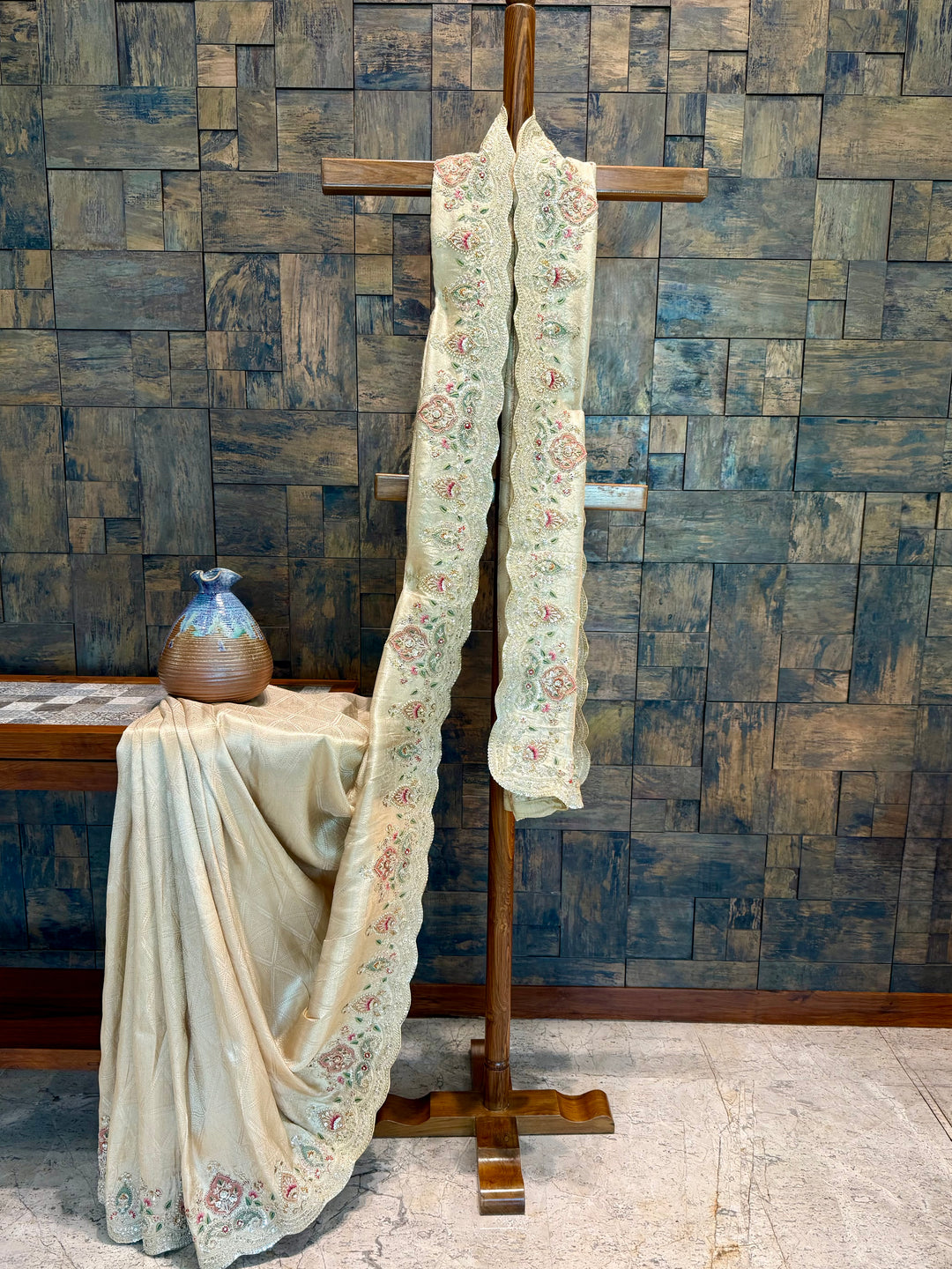 Off white Kanjivaram tissue silk saree with multicolor thread embroidery and floral paisley designs on scalloped borders. Decorative fabric draped over a wooden cross against a textured wall.