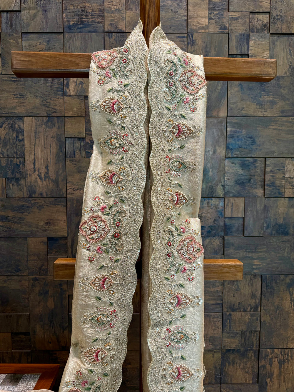 Detailed view of metallic thread hand embroidery in paisley and floral motifs on tissue silk base. Decorative fabric with intricate patterns hanging on a wooden cross against a wooden paneled wall.