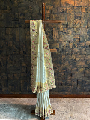Off White Kanjivaram Tissue Saree with embroidered peacock motifs and floral zari highlights displayed on a wooden stand.