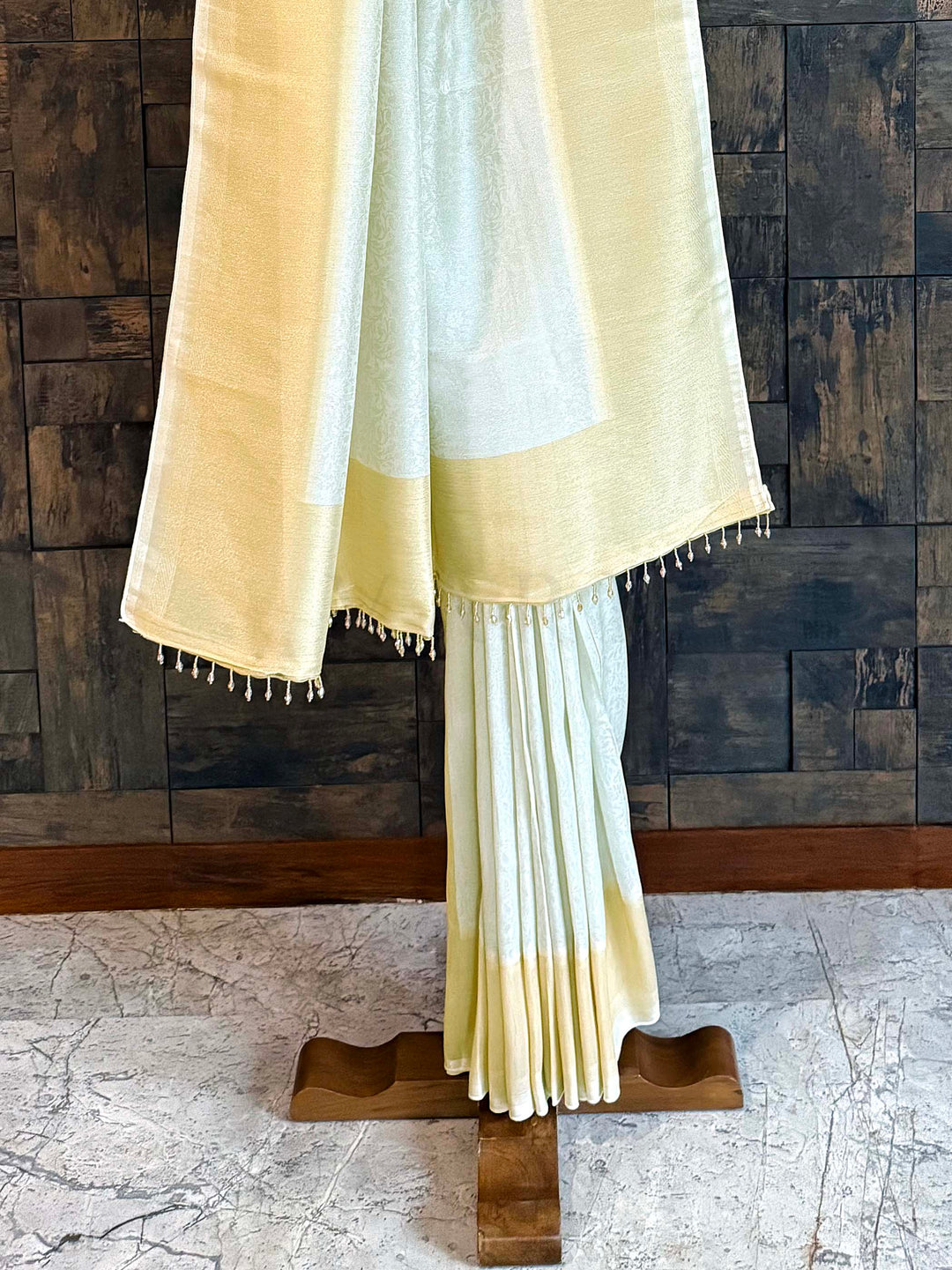 Beige and white saree draped over a wooden stand against a wooden paneled wall. Detailed view of floral creeper jaal woven on pistachio-mint tissue silk fabric.
