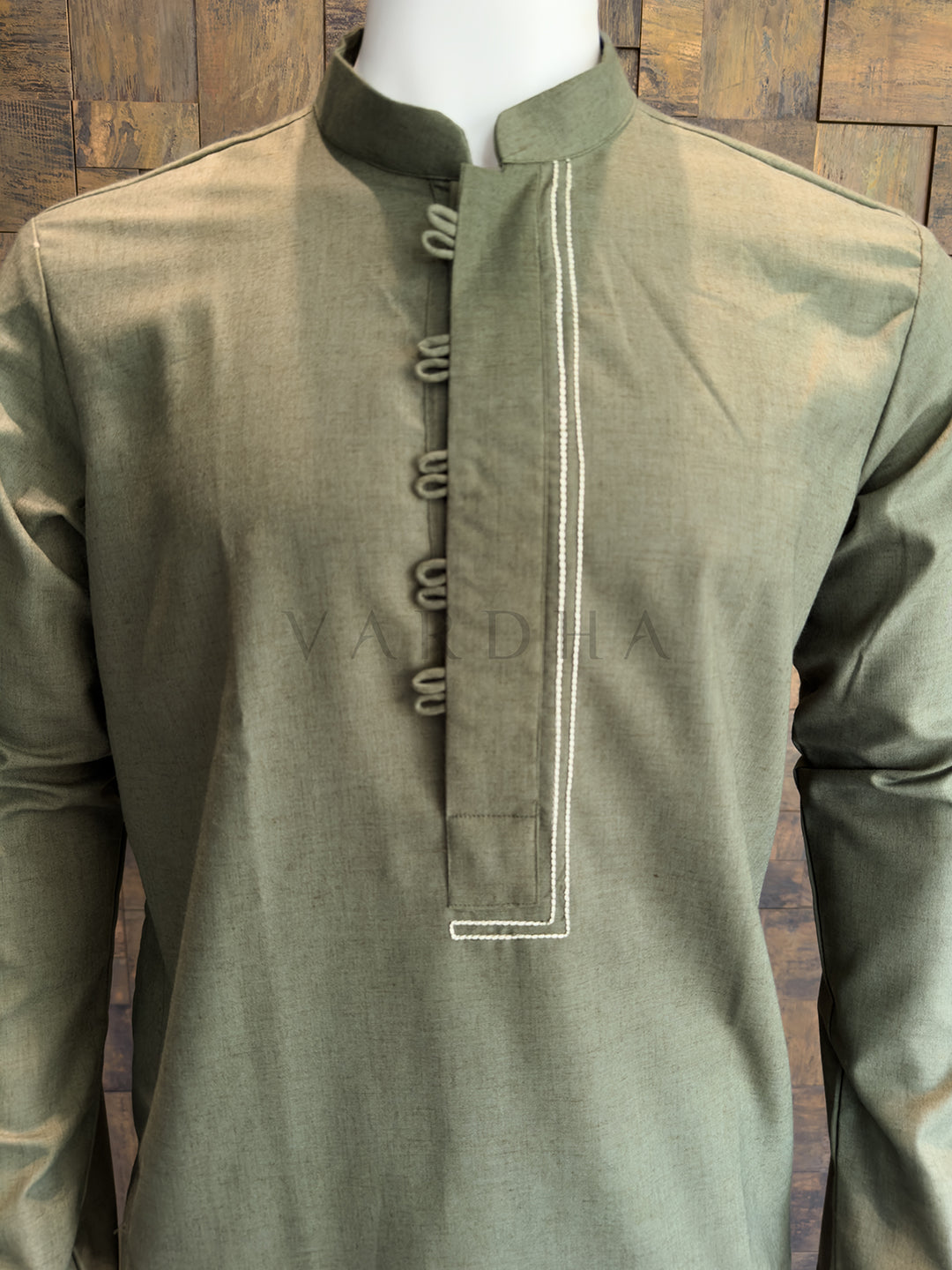 Green kurta with white embroidery on a wooden background Detailed view of the contrast white double stitching and fabric button loops on the olive kurta.