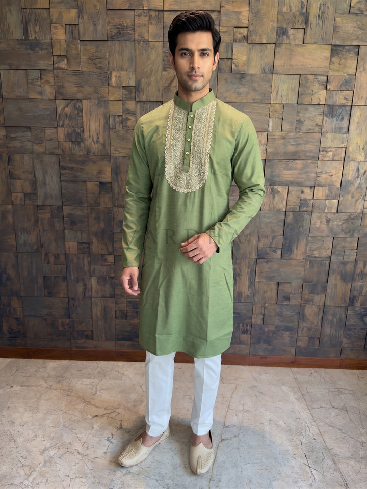 Man wearing a green kurta with white pants against a wooden paneled wall. Men’s Olive Green cotton kurta featuring a spectacular U-shaped chest yoke with heavy gold Zari threadwork, sparkling sequins, and a delicate pearl-beaded border.