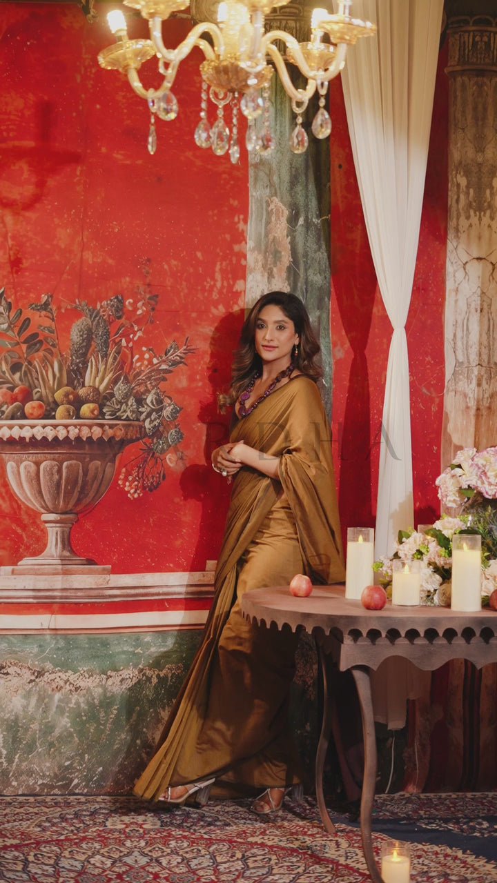 A woman wearing a flowing olive-brown satin saree stands in an ornate room with red patterned wallpaper, a chandelier, and a small table with flowers and candles. Beautiful woman posing gracefully in a soft olive green mal cotton saree, perfect for summer wear and daily office use.