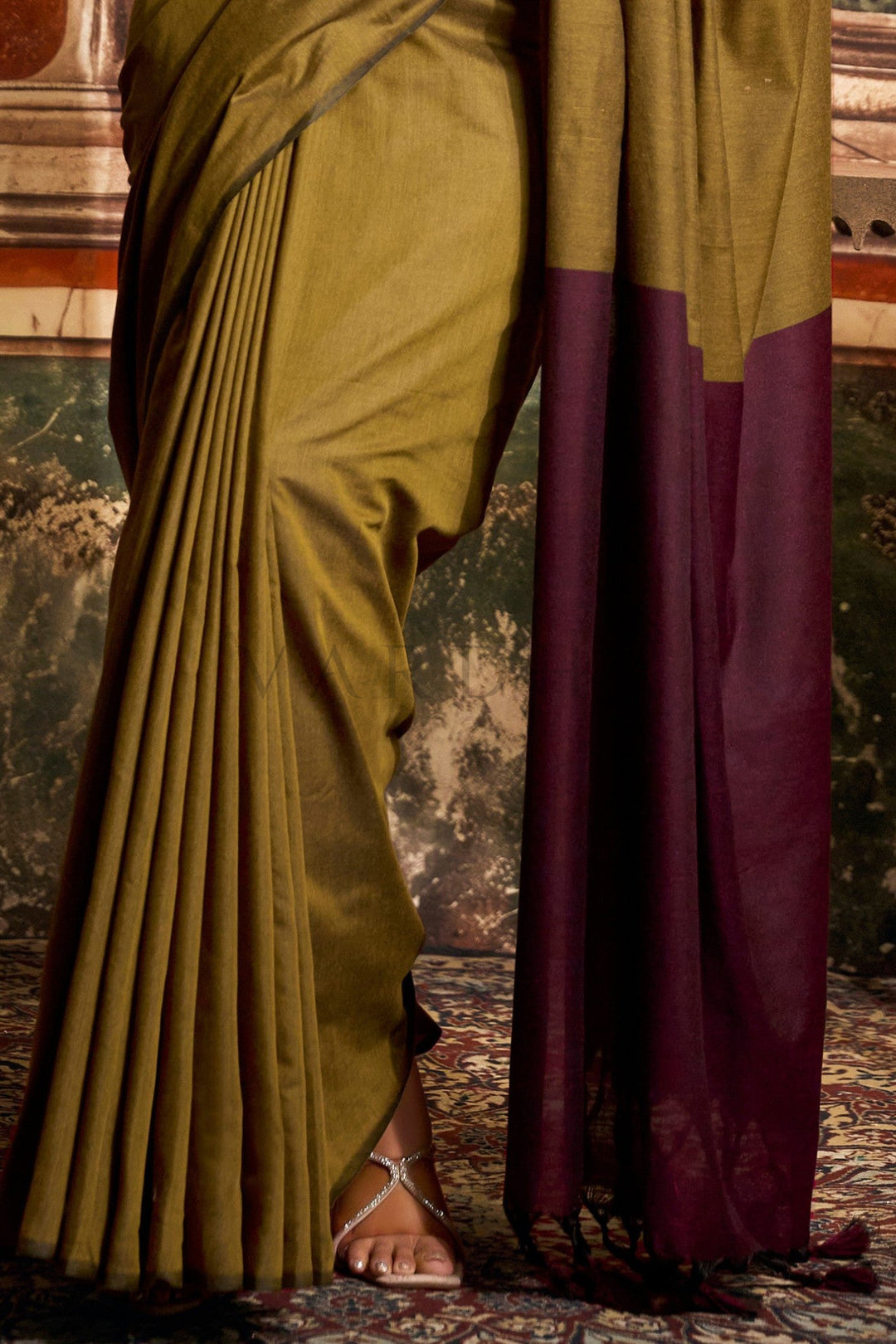 Person wearing a green and purple saree with a textured wall background Detailed fabric close-up of the olive green mal cotton saree highlighting the perfectly pleated texture and dark wine contrast pallu edge.