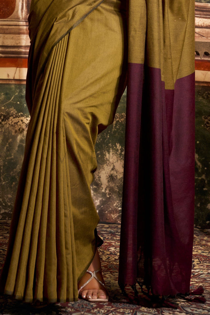 Person wearing a green and purple saree with a textured wall background Detailed fabric close-up of the olive green mal cotton saree highlighting the perfectly pleated texture and dark wine contrast pallu edge.