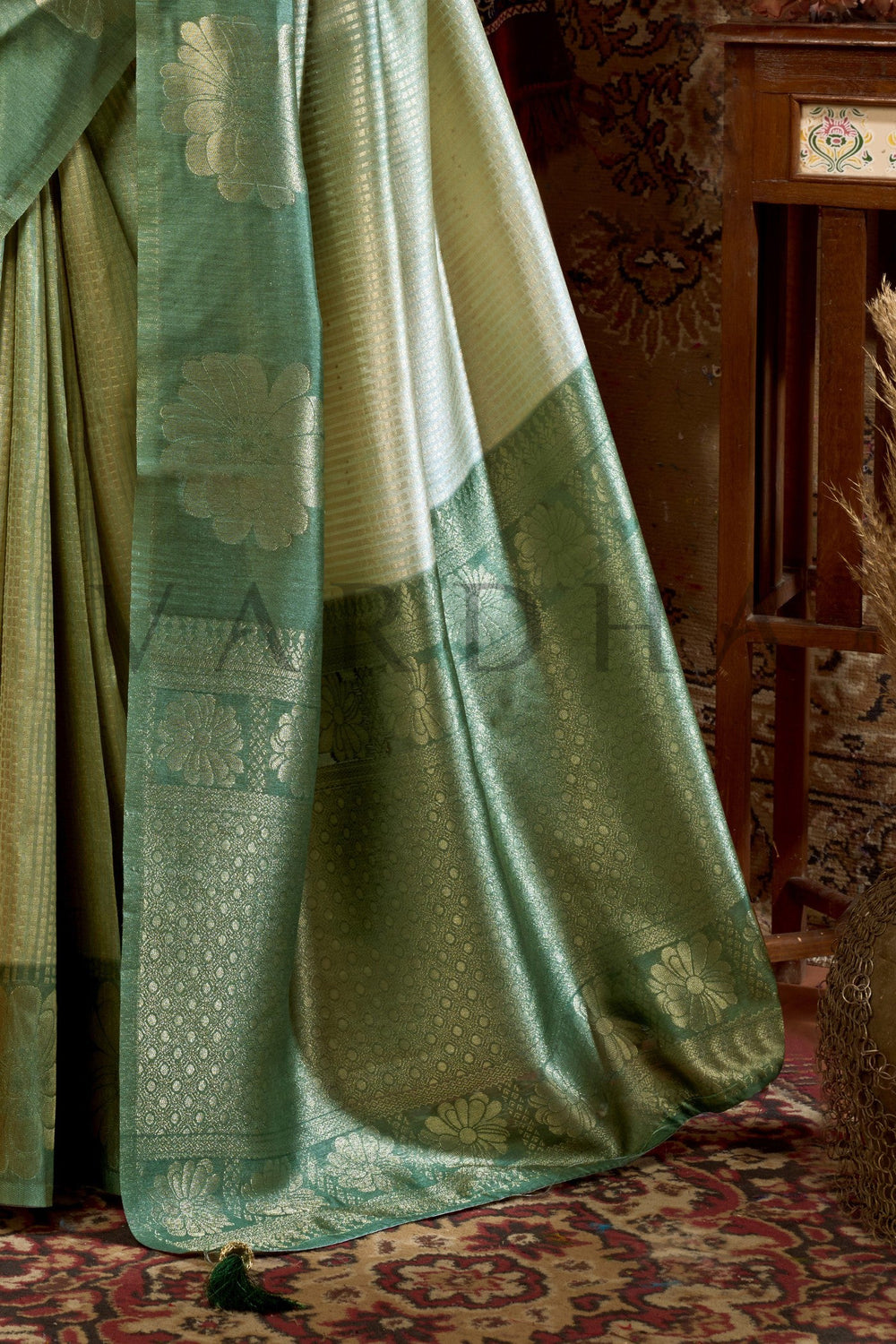 Green and beige fabric draped over a chair with a patterned rug underneath Close-up detail of the woven floral zari pallu and fancy jhumka tassels on the Olive Green Kota Silk Saree.
