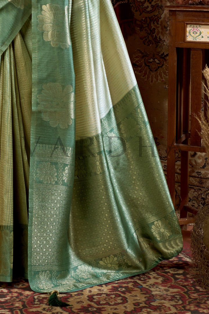 Green and beige fabric draped over a chair with a patterned rug underneath Close-up detail of the woven floral zari pallu and fancy jhumka tassels on the Olive Green Kota Silk Saree.