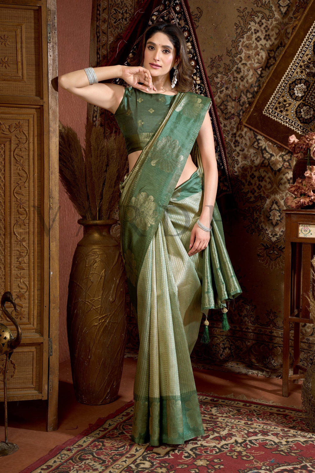 Woman in a green saree standing in an ornate room with patterned walls and carpet. Side profile highlighting the geometric woven body and rich olive green contrast border of the designer Kota silk saree.