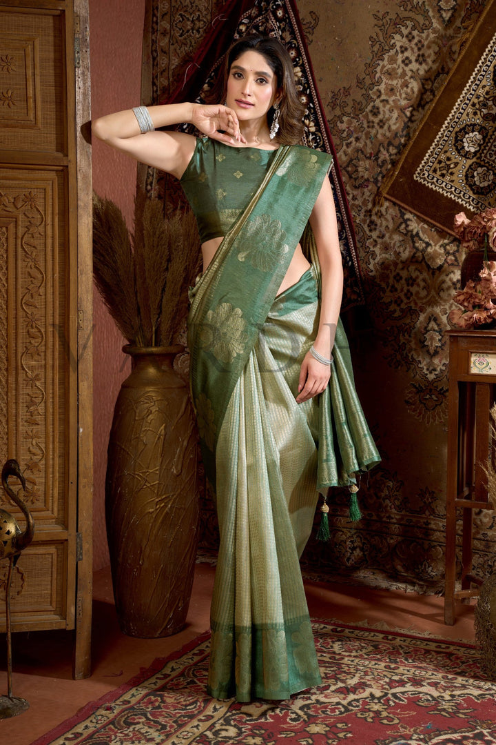 Woman in a green saree standing in an ornate room with patterned walls and carpet. Side profile highlighting the geometric woven body and rich olive green contrast border of the designer Kota silk saree.