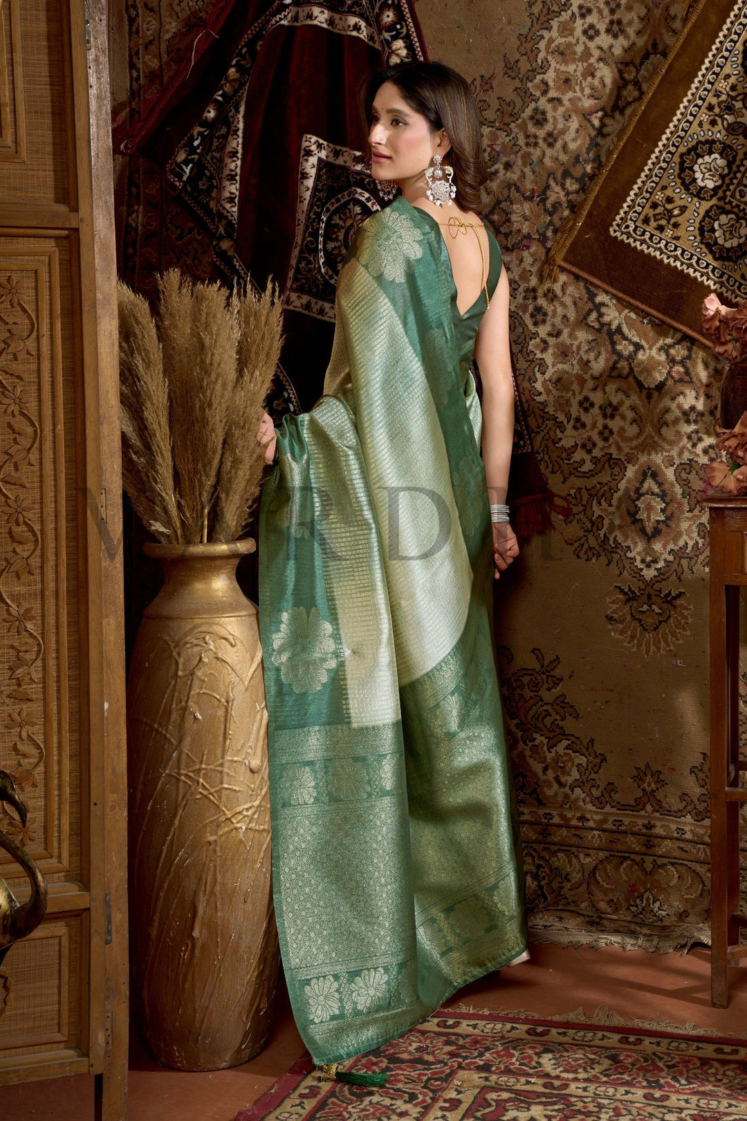 Woman holding a green saree in an ornate room with decorative wall and vase. Back view of the Olive Green Prijam Kota Silk Saree showcasing its flawless fall, soft drape, and intricate pallu design.