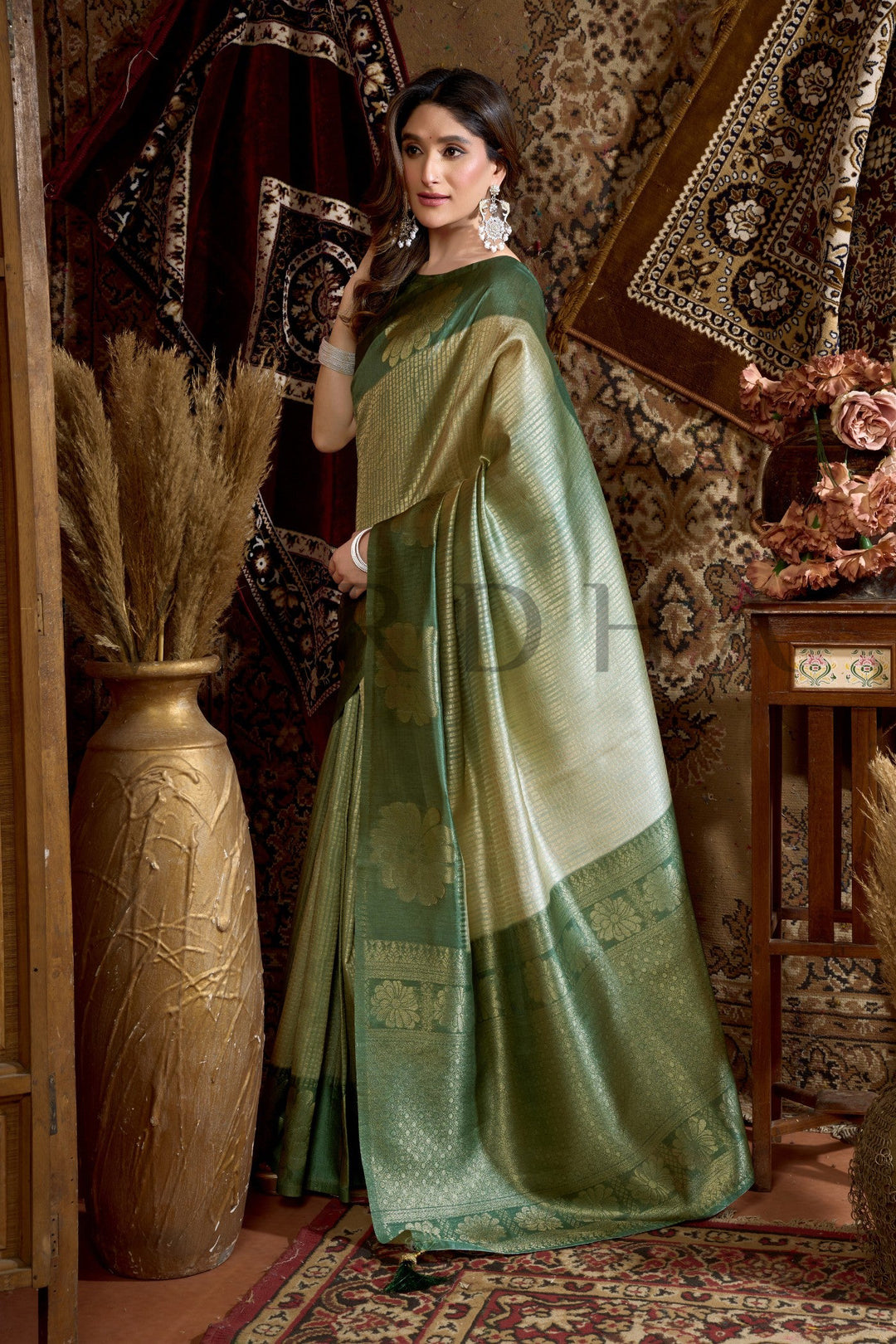 Woman in a green saree standing in an ornate room with decorative elements. Stylish pose featuring the dual-tone Olive Green and Golden Beige traditional Indian saree from House of Vardha.