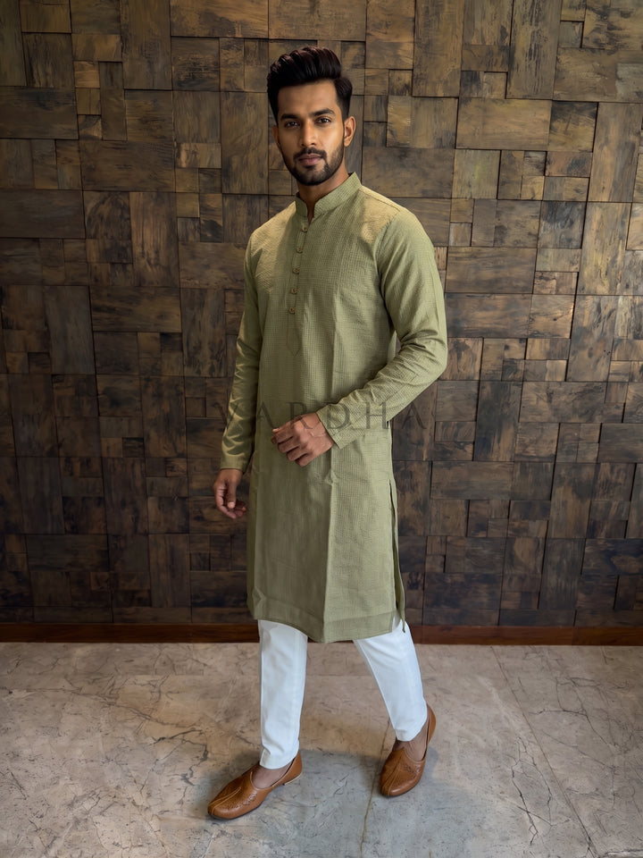 Man wearing a green kurta with white pants against a wooden paneled wall. Olive Green cotton kurta for men featuring a distinct woven micro-check texture and an extended chest placket with traditional button loops.