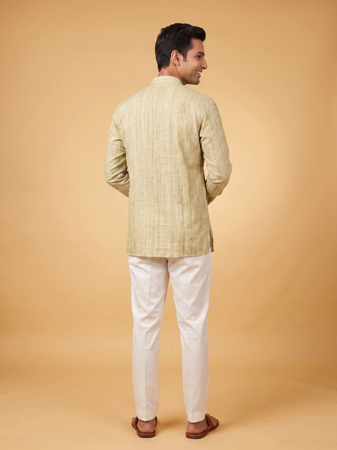 A man shown from the back wearing a light beige textured kurta and white trousers, standing against a solid warm beige studio background. Side-angle full-length view of a man walking in an olive green textured cotton short kurta and white trousers.