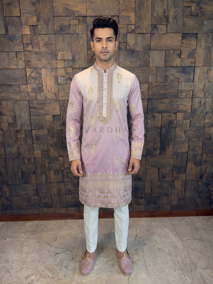 Man wearing a traditional embroidered kurta with a wooden paneled wall background. kurta features a captivating dual-tone fade from soft cream at the shoulders to a rich dusty mauve at the hem. It is exquisitely embellished with scattered gold zari floral motifs across the body. 