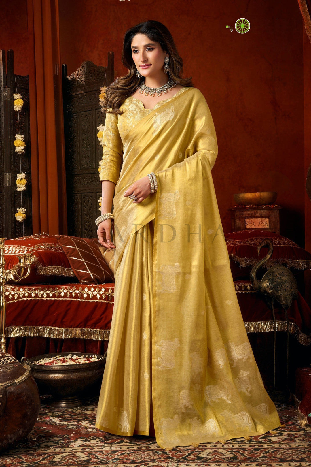 Woman in a yellow saree standing in an ornate room with red walls and decorative elements. Full front view of an elegant pale yellow heavy tissue saree featuring traditional silver woven cow buttas and a gold zari border.