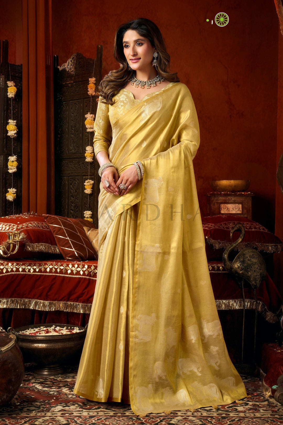A woman wearing a flowing golden-yellow saree with subtle floral motifs, standing in a richly decorated traditional room with red walls and ornate furniture. Beautiful woman standing gracefully in a light gold tissue silk saree, showcasing the luxurious drape and matching woven blouse.