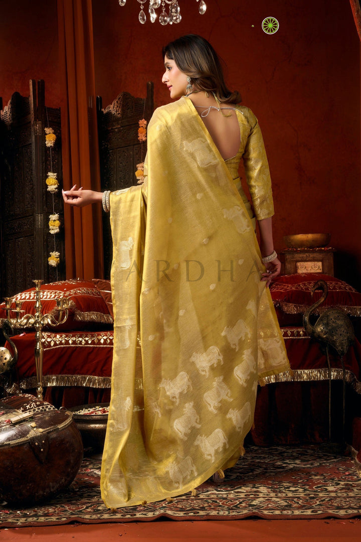 Woman in a yellow saree with a floral pattern in a traditional setting. Back view of the radiant pale yellow tissue saree, emphasizing the rich metallic sheen, woven silver cow designs, and elegant fancy tassels.