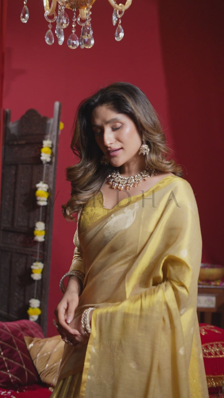 A woman wearing a golden yellow saree and matching blouse with ornate jewelry, standing in a richly decorated room with red walls and a chandelier. Glamorous evening shot of a woman looking down gracefully while wearing the beautiful light gold heavy tissue saree, highlighting its regal metallic glow.