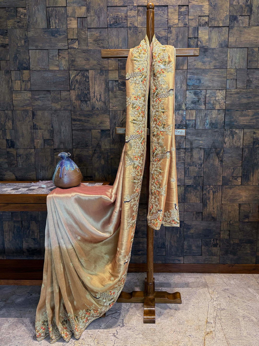 Peach Pink Kanjivaram Tissue Silk Saree draped with scalloped hand-embroidered border and golden sheen. Decorative fabric draped over a stand against a textured wall.