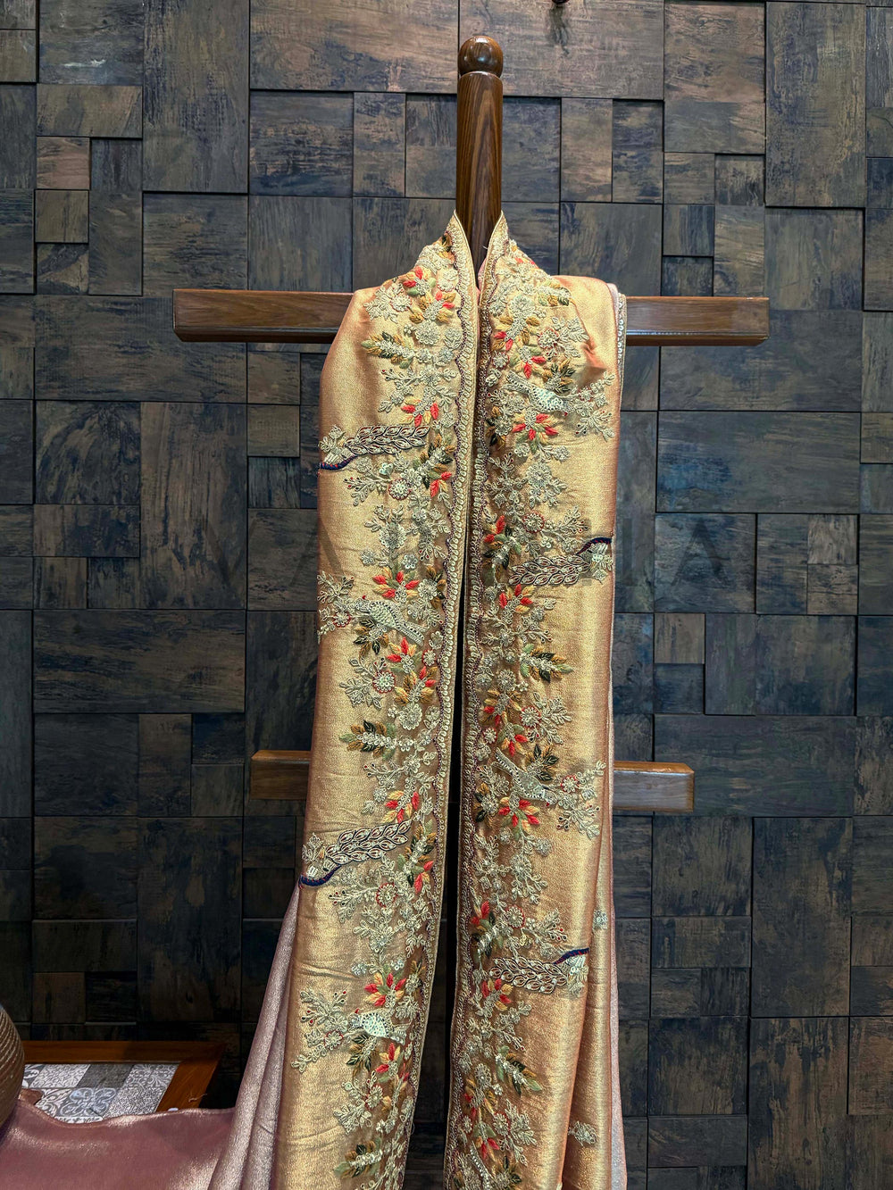 Close-up of scalloped border with coral and ivory florals, sequin accents, and antique zardozi threads. Decorative fabric with floral patterns on a wooden hanger against a textured wall.