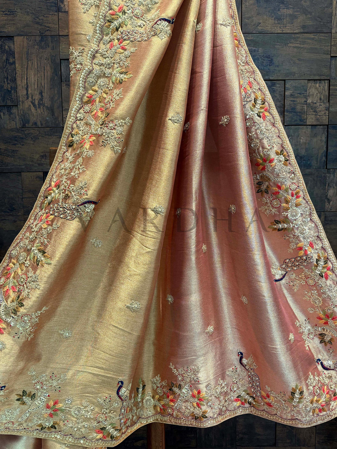Pallu detailing showcasing intricate floral threadwork and peacock zari motifs on pink-beige base. Decorative fabric with intricate embroidery on a wooden background