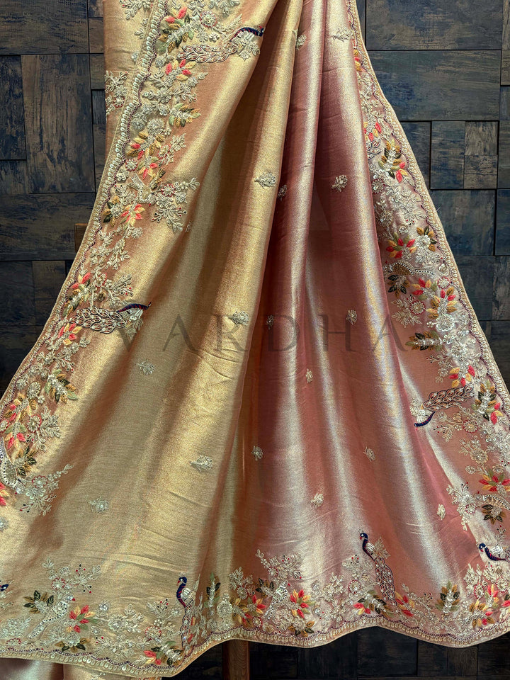 Pallu detailing showcasing intricate floral threadwork and peacock zari motifs on pink-beige base. Decorative fabric with intricate embroidery on a wooden background