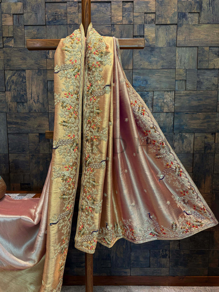 Border close-up with navy thread peacocks, floral vines, and resham detailing in multi-color. Decorative Saree with intricate patterns draped over a wooden cross against a wooden paneled wall.