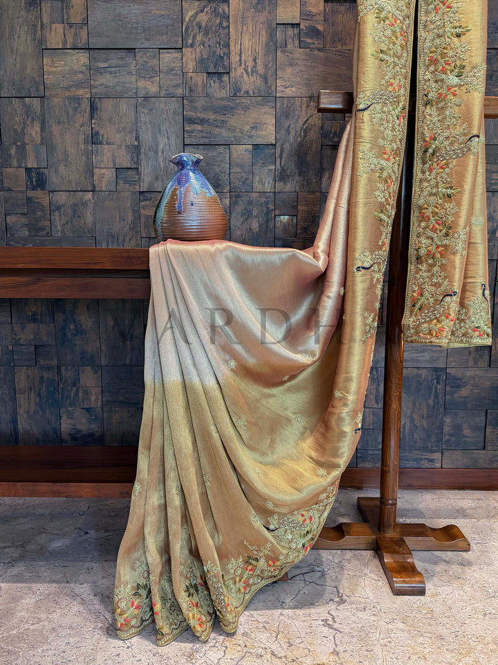 Full edge view of saree showing metallic dual-tone transition between peach, pink, and golden hues. Decorative fabric draped over a vase against a textured wall.
