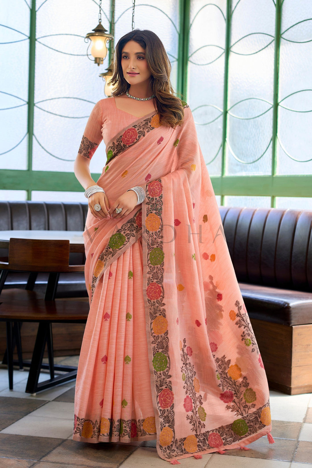 A woman wearing a peach-pink georgette saree with colorful floral prints and a matching printed blouse, standing in a restaurant setting. Full-length view of a woman wearing an elegant peach linen jacquard handloom saree with a multi-colored floral woven border and fancy tassels.