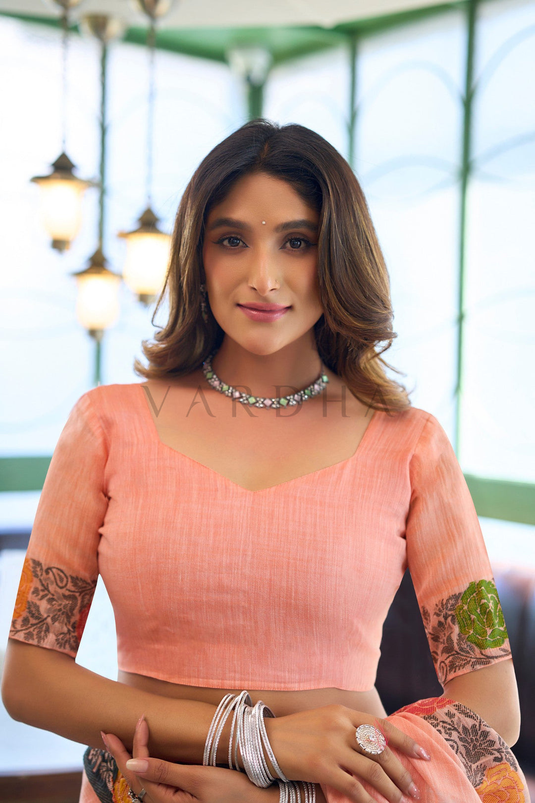 A woman wearing a peach-colored sleeveless blouse with embroidered patterns on the sleeves, paired with a silver beaded necklace and stacked silver bangles, set against a softly blurred background with hanging lights. Close-up portrait of the matching peach blouse featuring a v-neckline and woven floral jacquard borders on the sleeves.