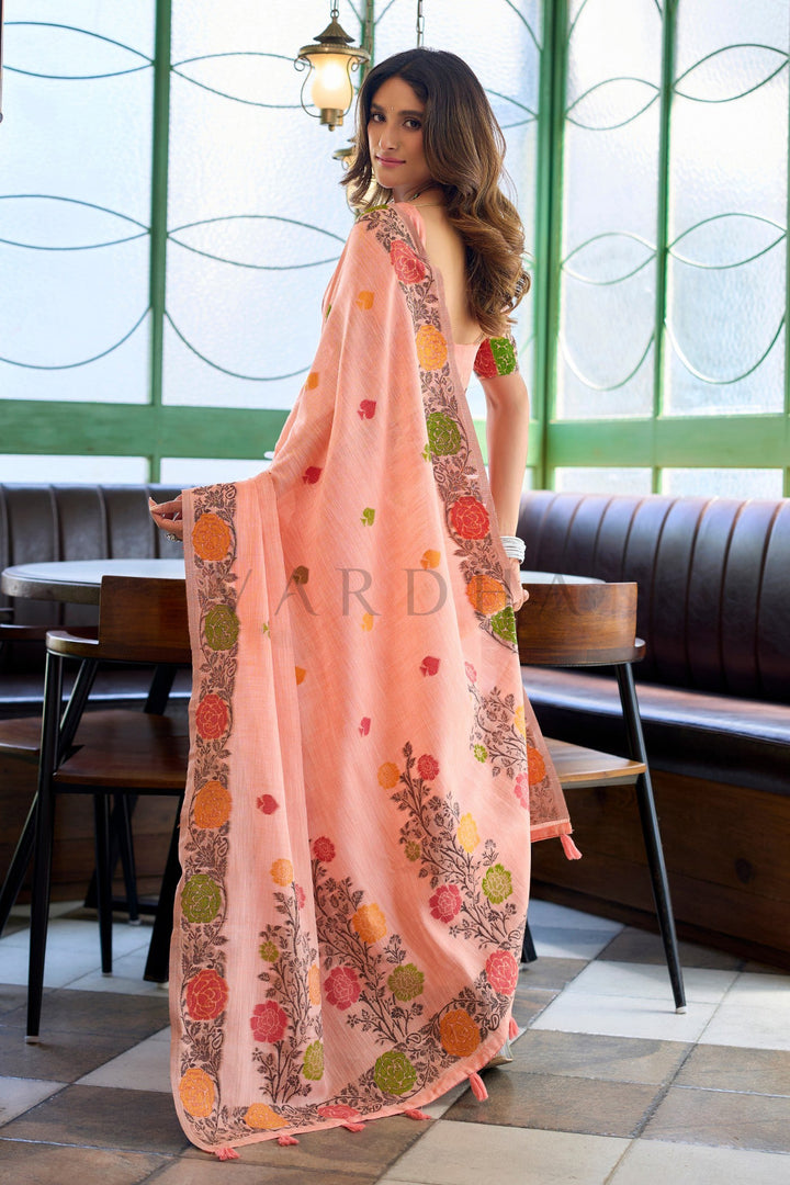 A woman wearing a peach-colored sleeveless maxi dress with colorful floral patterns and a matching dupatta, standing in a cafe setting. Back view of the peach linen jacquard saree displaying the stunning pallu design with large woven roses and fancy pink tassels at the edge.