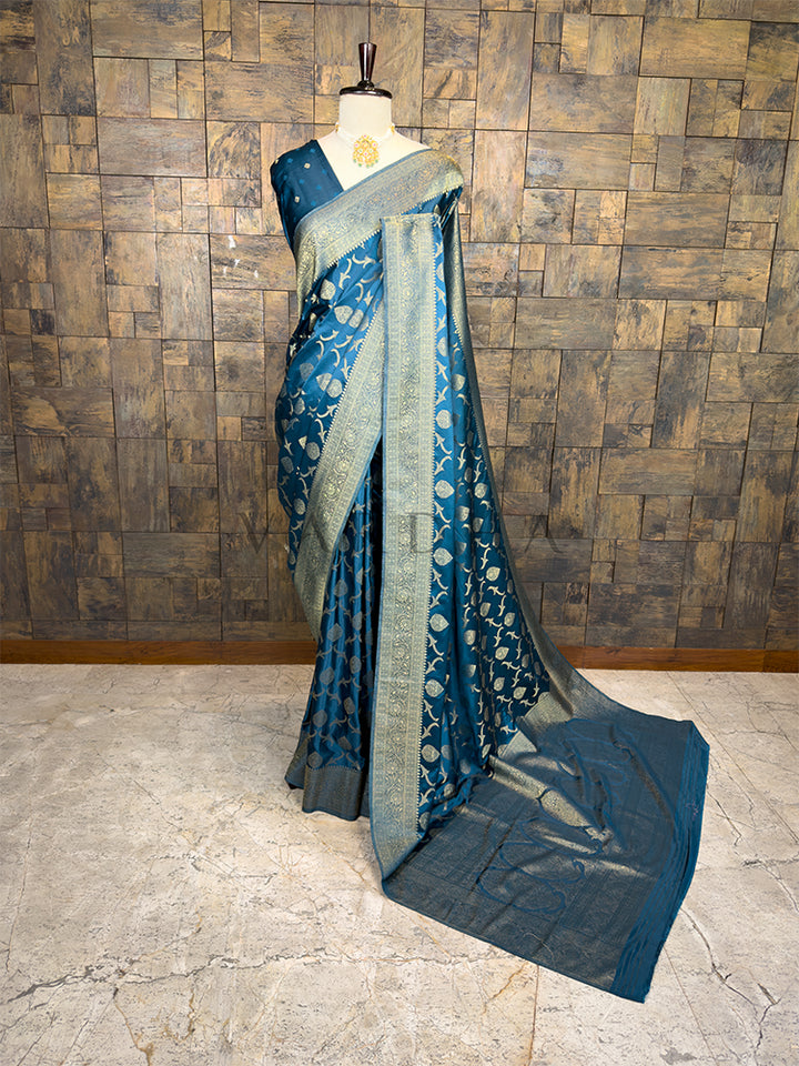Blue saree with a pattern on a mannequin against a wooden paneled wall. House of Vardha Peacock Blue Golden Zari Banarasi Satin Silk Saree featuring heavy traditional woven leaf motifs, elegantly draped on a mannequin. We offer Ready to wear, Pre drape service, Saree Add on, Custom Stitched Blouse, Fall and Edging, Gift Packaging at Complimentary Price!