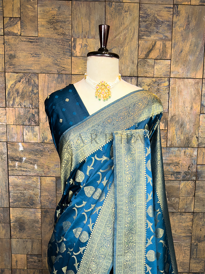 Blue saree with gold patterns on a mannequin against a wooden paneled wall. Blue saree with gold patterns on a mannequin against a wooden paneled wall. Lustrous Peacock Blue Golden Zari Banarasi Satin Silk Saree by House of Vardha showcasing intricate golden zari craftsmanship and a rich matching border. We offer Ready to wear, Pre drape service, Saree Add on, Custom Stitched Blouse, Fall and Edging, Gift Packaging at Complimentary Price!