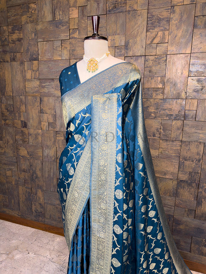 Blue saree with white patterns on a mannequin against a wooden paneled wall. Blue saree with white patterns on a mannequin against a wooden paneled wall. Close-up view of the House of Vardha Peacock Blue Golden Zari Banarasi Satin Silk Saree highlighting the detailed golden zari leaf patterns on the pleats and body. We offer Ready to wear, Pre drape service, Saree Add on, Custom Stitched Blouse, Fall and Edging, Gift Packaging at Complimentary Price!
