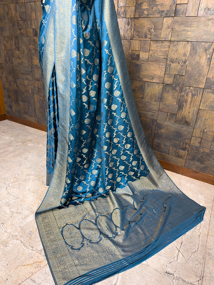 Blue and gray saree draped over a wooden surface. Blue and gray saree draped over a wooden surface. Elegant Peacock Blue festive Banarasi Satin Silk Saree from House of Vardha with a radiant golden zari weave, perfect for evening wear and special occasions. We offer Ready to wear, Pre drape service, Saree Add on, Custom Stitched Blouse, Fall and Edging, Gift Packaging at Complimentary Price!