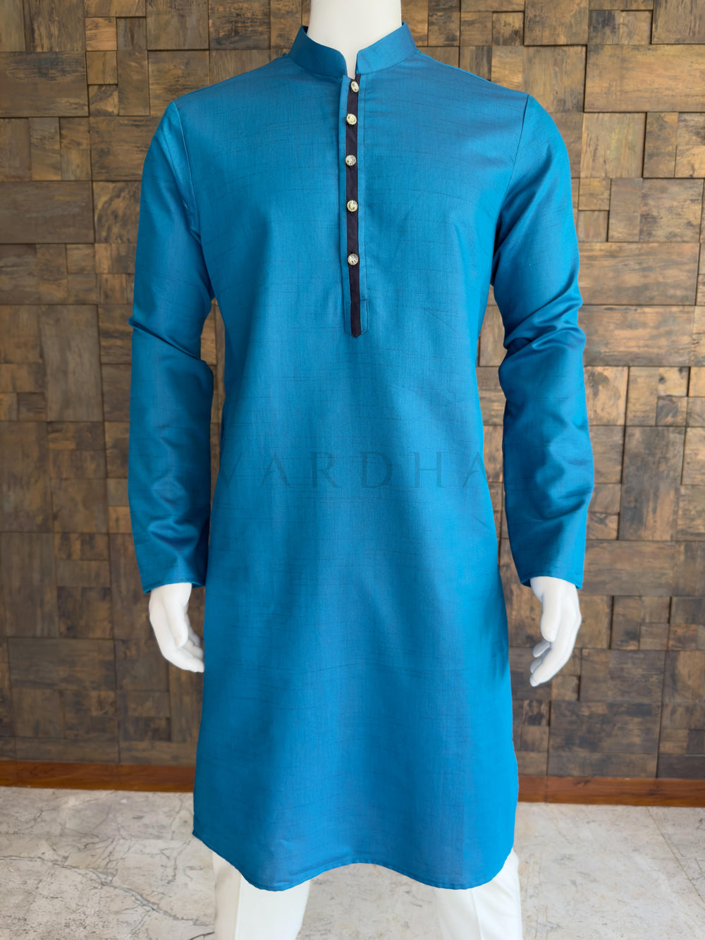 Blue embroidered kurta on a mannequin against a wooden paneled wall. Close-up of the teal blue men's festive kurta showcasing the fine slub cotton texture and a striking dark navy center placket with premium jeweled decorative buttons.