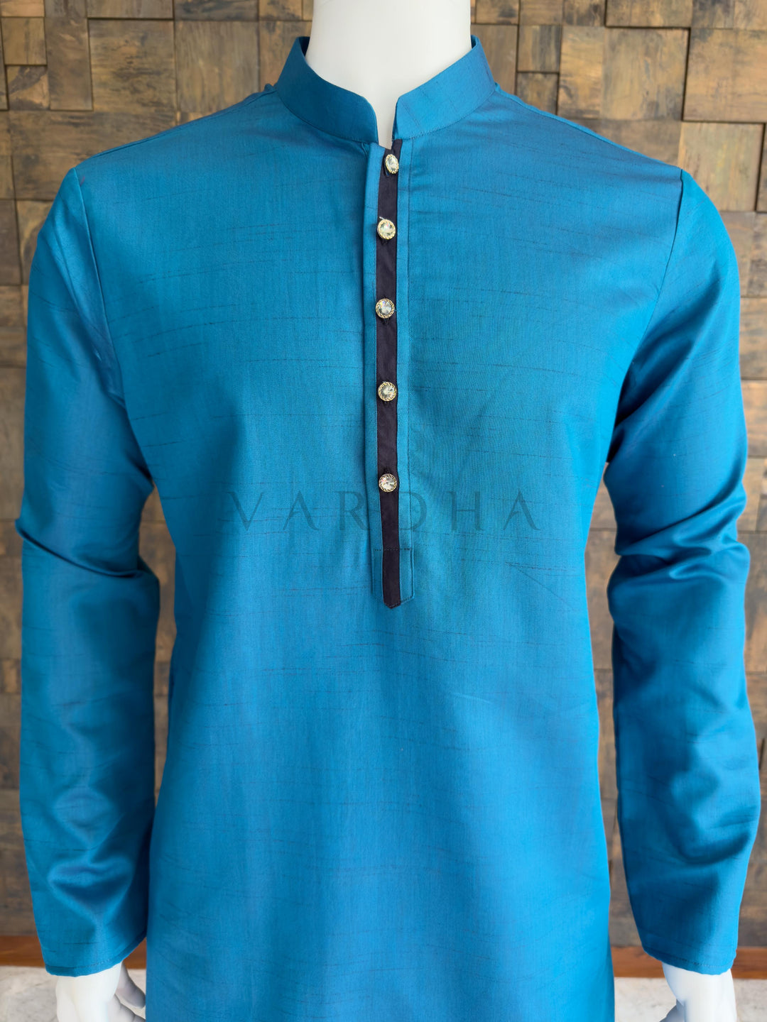 Blue embroidered shirt on a mannequin against a wooden paneled wall. Detailed chest view highlighting the crisp mandarin collar, jeweled buttons, and contrast dark placket on the premium teal blue textured cotton designer kurta for men.