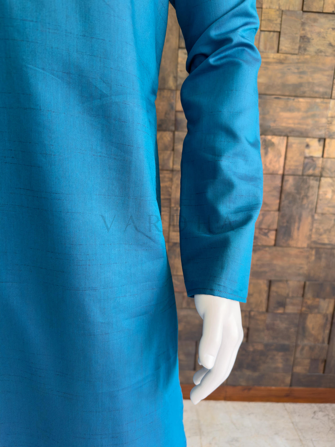 Blue embroidered garment on a mannequin against a wooden paneled wall. Close-up of the full sleeve and side drape of the vibrant teal blue textured cotton kurta, showcasing the breathable premium fabric and clean, elegant minimalist aesthetic.