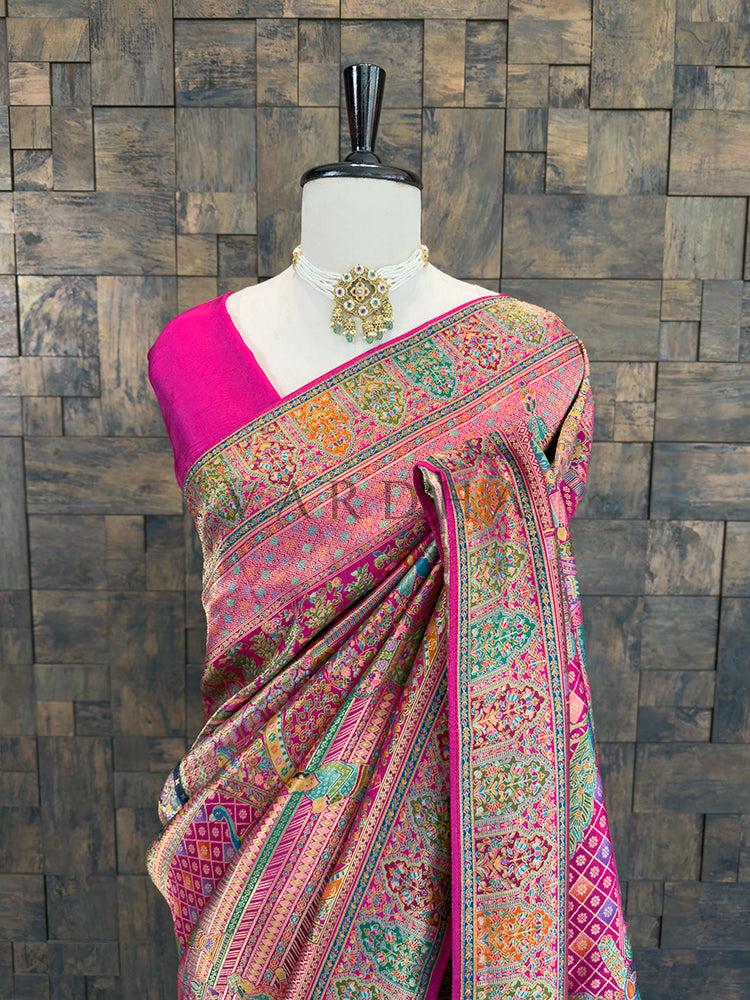 Saree with intricate patterns on a mannequin against a wooden paneled wall. Close-up of a bright pink traditional bridal silk saree draped over the shoulder, highlighting vibrant woven floral and geometric patterns in multi-color Meenakari threadwork, elegantly accessorized with a pearl choker.