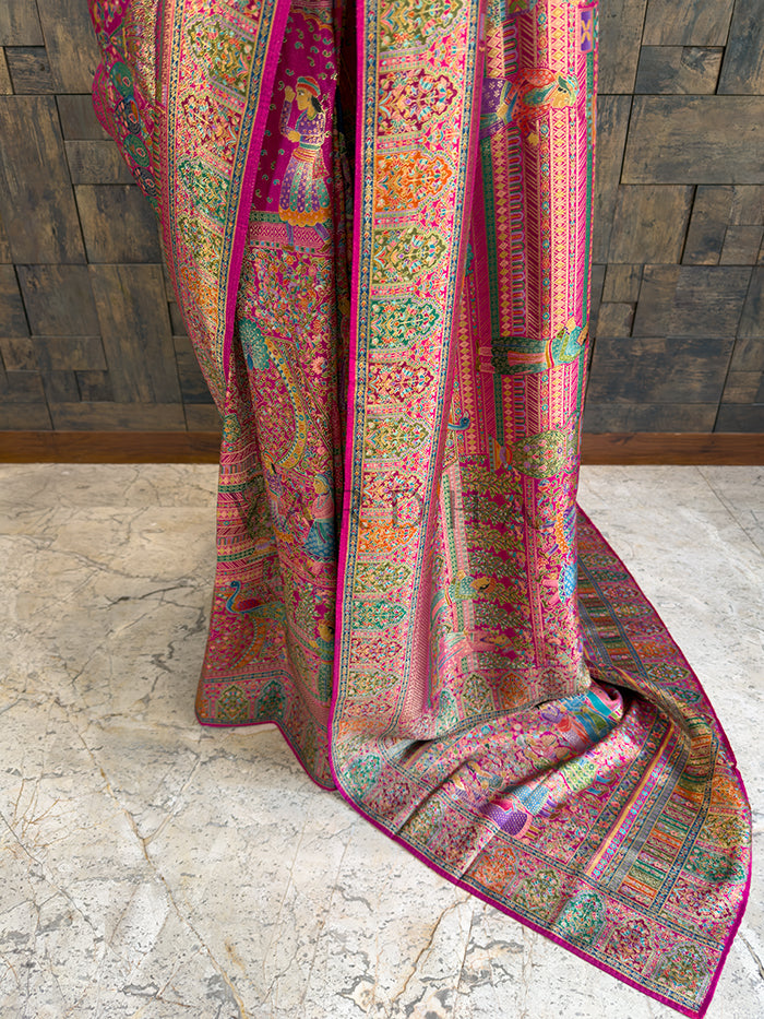 Colorful patterned fabric draped over a surface with a wooden panel background Lower drape of a heavy heritage woven Meenakari silk saree, showcasing the rich pink base and elaborate multi-colored borders with intricate floral vines and traditional human figure motifs cascading toward the floor.