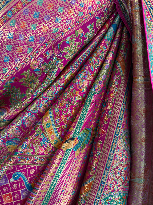 Colorful patterned fabric with intricate designs in pink, green, and blue. Macro texture shot of a premium pink Meenakari silk saree in gathered pleats, detailing the vibrant multi-colored woven diamond lattice, traditional Indian bird and figure motifs, and rich heritage weaving techniques.