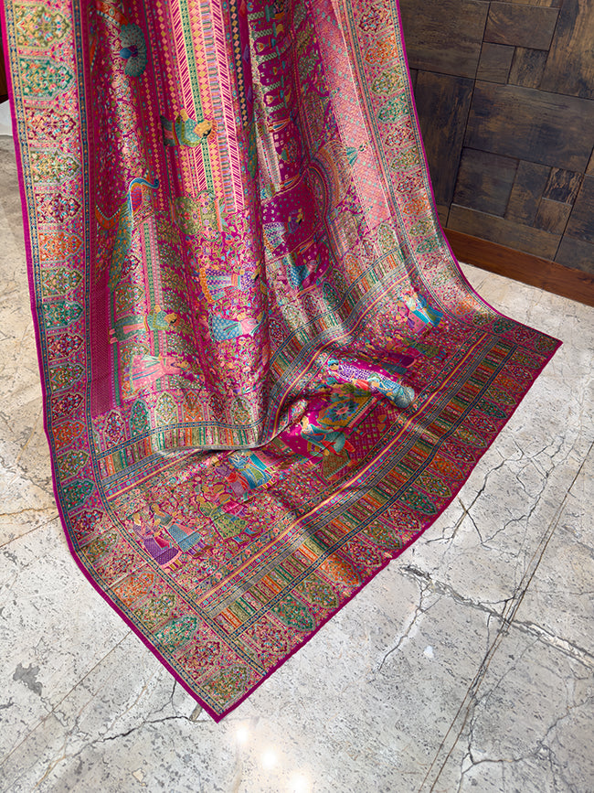 Colorful patterned fabric draped over a wooden surface Trailing edge of a stunning magenta pink festive saree displayed on a stone floor, revealing a heavily woven pallu with intricate royal court figures, traditional Indian motifs, and elaborate multi-colored borders.